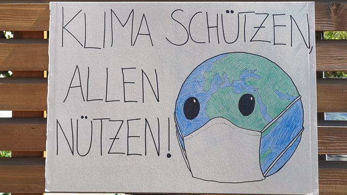 Ein selbstgebasteltes Plakat mit der Aufschrift "Klima schützen, allen nützen" hängt an einem Bretterzaun.